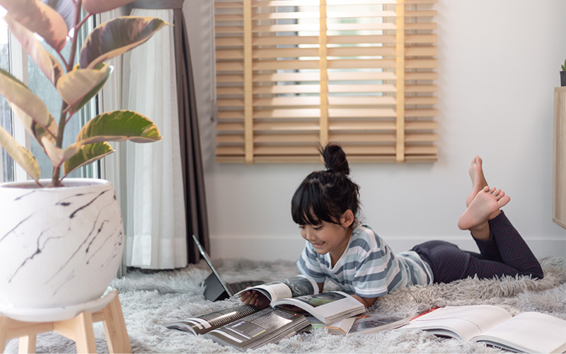 Child lying on carpet reading with books and tablet in a sunlit room with plant and curtains.