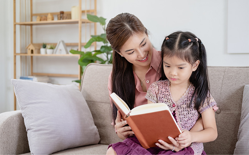 Asian woman reading a book with a child on a beige sofa in a cozy living room with shelves and plants.