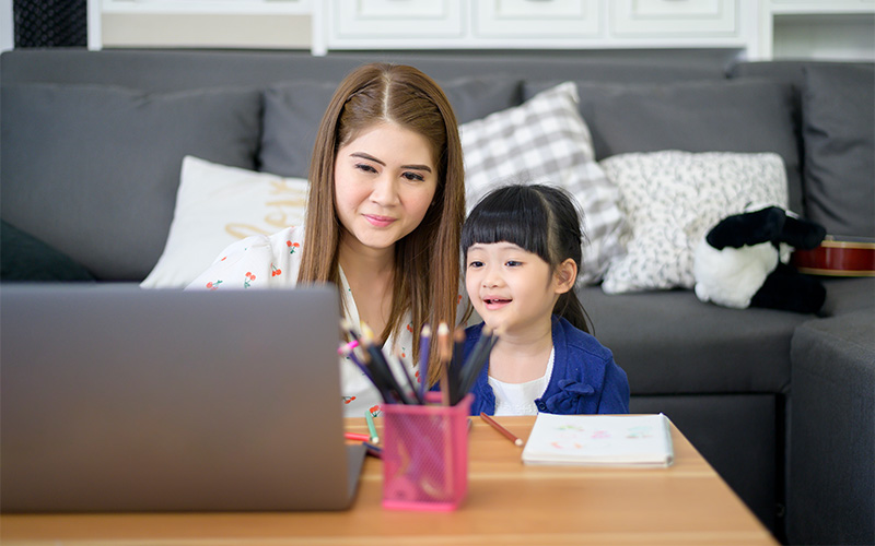 An adult and child sit at a table using a laptop, surrounded by art supplies in a cozy living room.