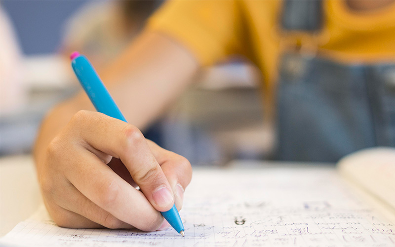 Close-up of a child’s hand holding a blue pen, writing in a notebook on a desk.