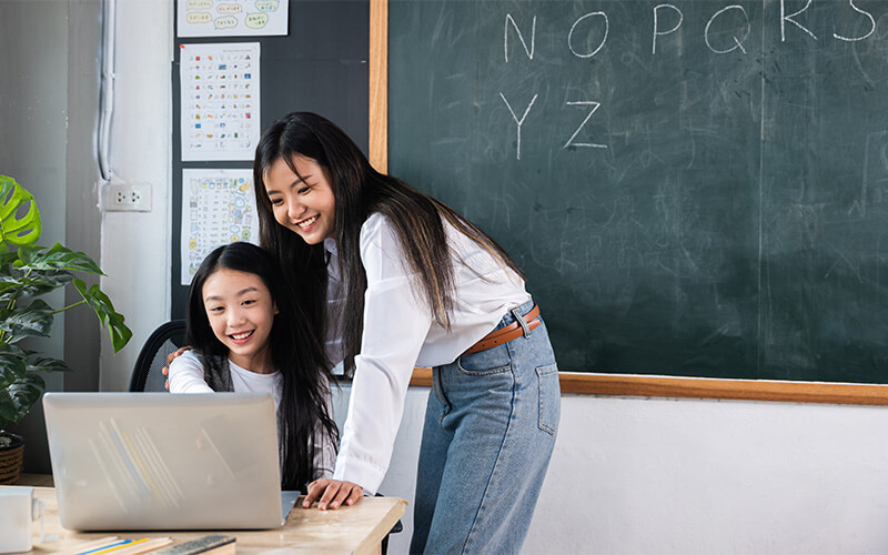 Teacher helping a young student use a laptop in a classroom.