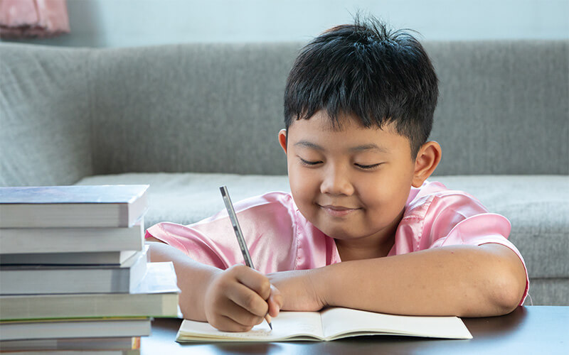 Young child smiling while writing in a notebook at a table with stacked books nearby.
