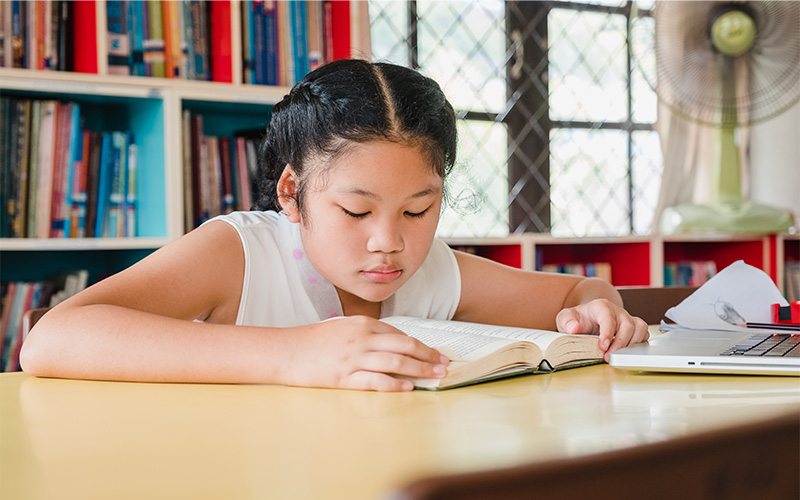 Young girl reading a book at a table in a library.